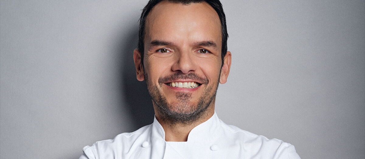 Portrait Steffen Henssler - in weißer Kochjacke vor einer grauen Rückwand