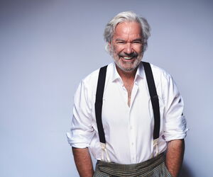 Stefan Gwildis in die Kamera lachend mit weißem Hemd und Hosenträgern vor blauem Hintergrund.