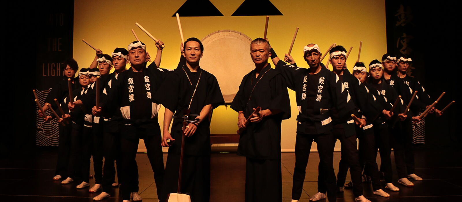 Kokubu Band mit Trommeln und Schlagstöckern in der Hand vor einem gelben Hintergrund