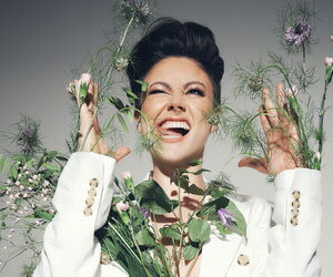 Tahnee mit einem Blumenstrauß im Arm, weißem Anzug in die Kamera lachend.
