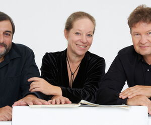 Die Musiker:innen des Trios auf einen Tisch gelehnt in die Kamera schauend