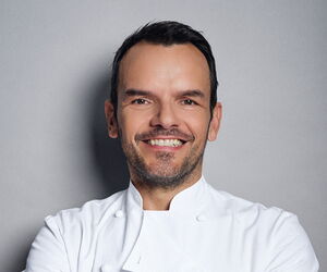 Portrait Steffen Henssler - in weißer Kochjacke vor einer grauen Rückwand