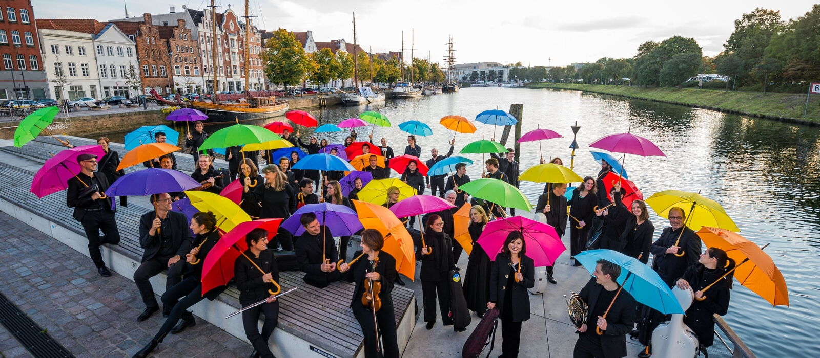 Orchestermitglieder auf dem Drehbrückenplatz stehend mit Instrumenten unter bunten Regenschirmen.