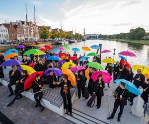 Orchestermitglieder auf dem Drehbrückenplatz stehend mit Instrumenten unter bunten Regenschirmen.
