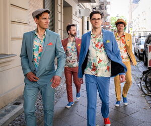 Bodo Wartke und die SchönenGutenA-Band in farbigen Anzügen mit bunten Hemden auf dem Bürgersteig laufend.