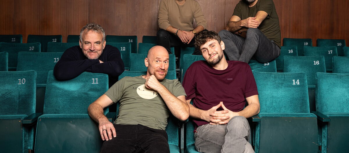 Die Bandmitglieder in einem Kino verteilt sitzend und in die Kamera schauend.