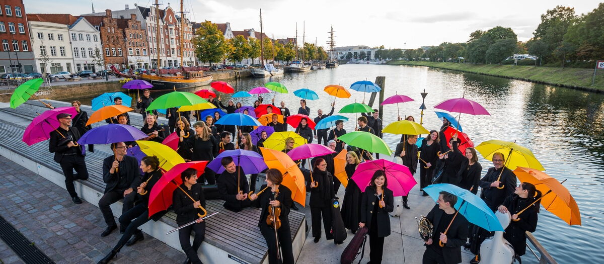 Orchestermitglieder auf dem Drehbrückenplatz stehend mit Instrumenten unter bunten Regenschirmen.