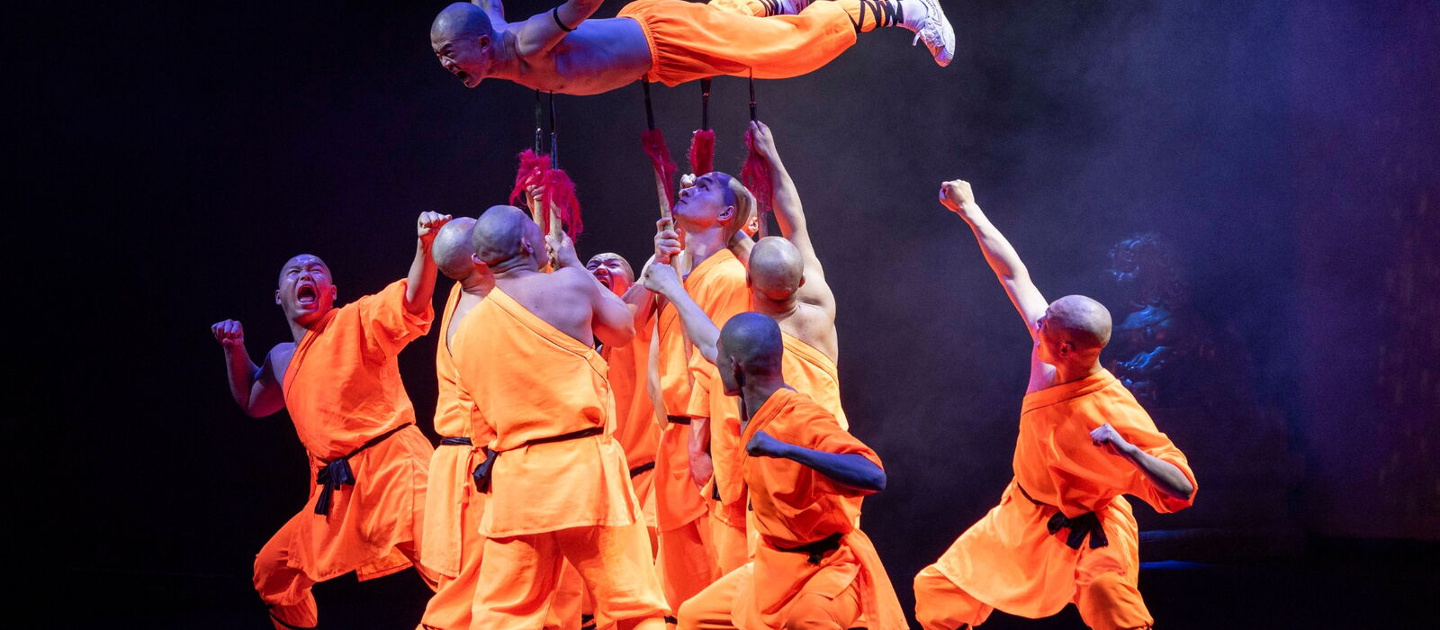 Shaolin Mönche in orangen Kostümen in Aktion auf einer Bühne.