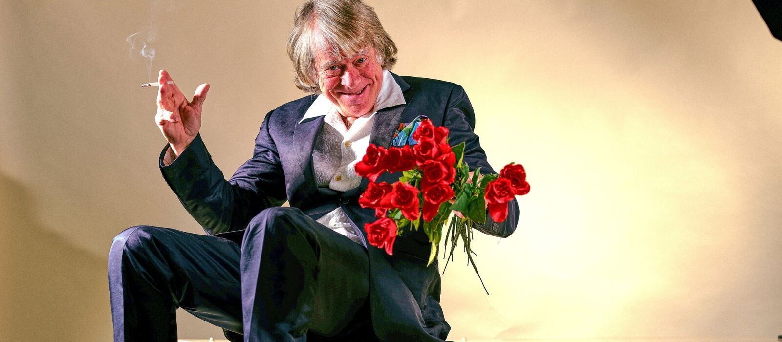 Helge Schneider auf einem Tisch sitzend mit Zigarette und roten Rosen in der Hand.