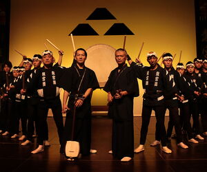 Kokubu Band mit Trommeln und Schlagstöckern in der Hand vor einem gelben Hintergrund