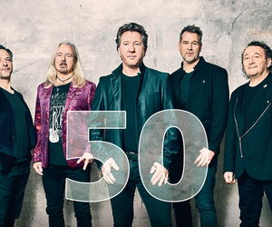 Die Bandmitglieder von Karat vor einer beigen Wand mit einem roten Karat-Schriftzug darüber und einer 50 vor den Bandmitgliedern.