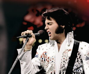 Elvis ins Mikrofon singend in weißem Anzug mit umhängender Gitarre.