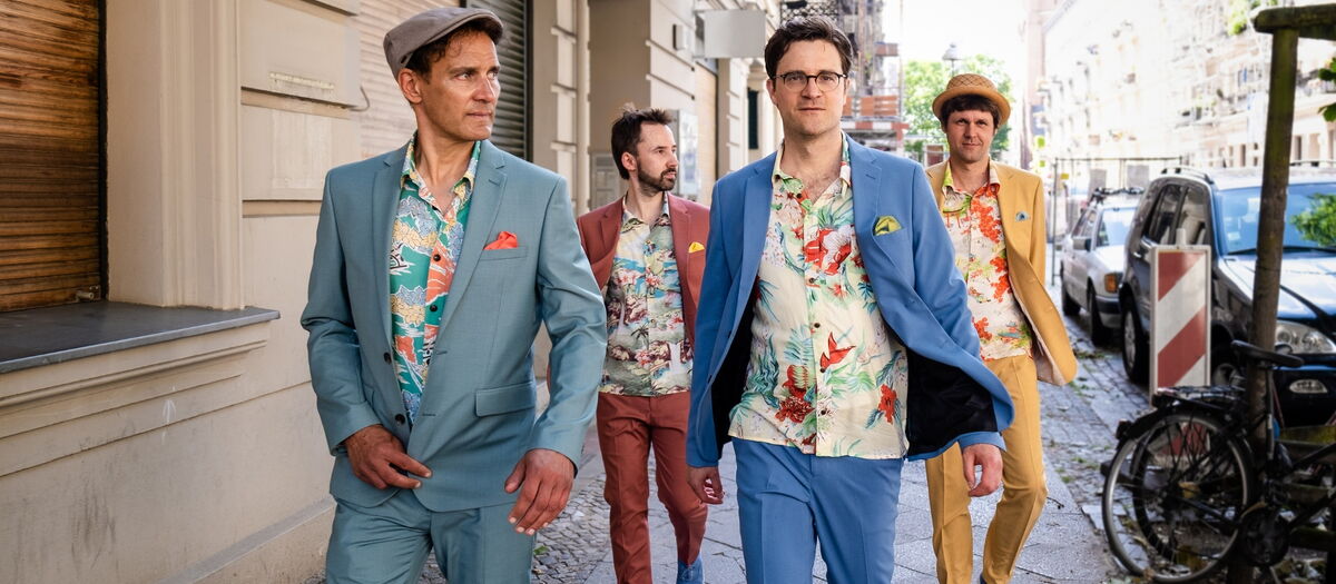 Bodo Wartke und die SchönenGutenA-Band in farbigen Anzügen mit bunten Hemden auf dem Bürgersteig laufend.