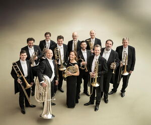 Das Blechbläser-Ensemble der Berliner Philharmoniker mit ihren Instrumenten vor einem blaugrünen Glasfenster stehend.