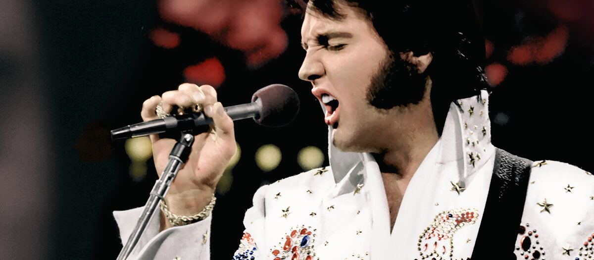 Elvis ins Mikrofon singend in weißem Anzug mit umhängender Gitarre.