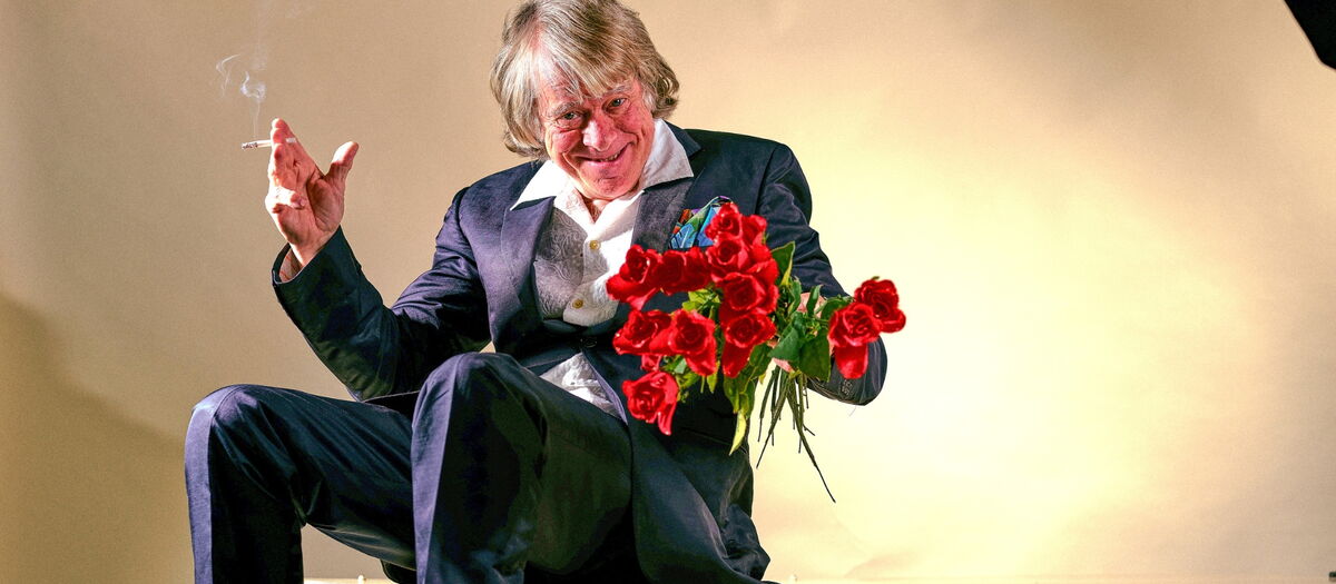 Helge Schneider auf einem Tisch sitzend mit Zigarette und roten Rosen in der Hand.