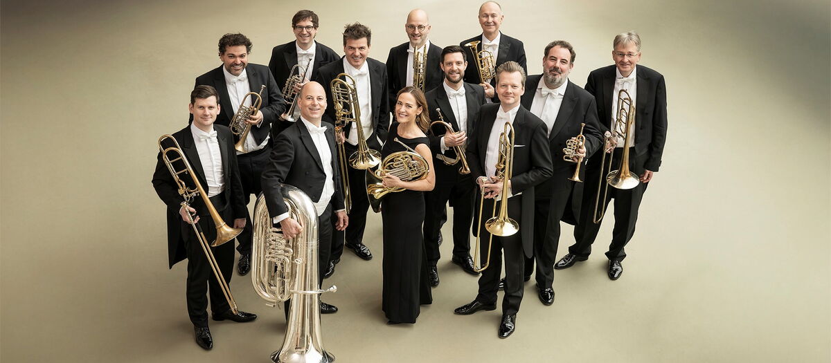 Das Blechbläser-Ensemble der Berliner Philharmoniker mit ihren Instrumenten vor einem blaugrünen Glasfenster stehend.